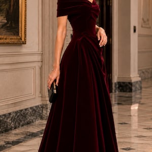 Puede incluir: Un vestido largo hasta el suelo de terciopelo color burdeos intenso con escote de hombros descubiertos. El dise&ntilde;o presenta un corpi&ntilde;o ajustado con fruncidos sutiles en la cintura y una falda amplia y fluida que llega al suelo. Se sostiene un peque&ntilde;o bolso de mano negro.