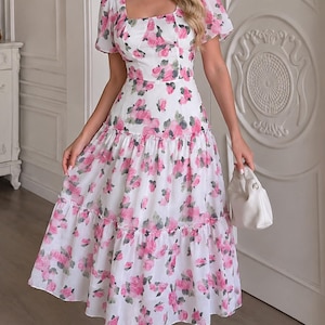 Può includere: Un abito midi bianco con stampa floreale di rose rosa, maniche corte a sbuffo, scollo quadrato e gonna a balze. L'abito è abbinato a sandali bianchi con cinturini e una piccola borsa bianca, creando un look coerente con accenti floreali.