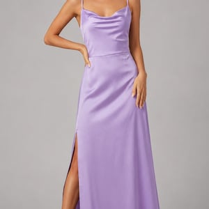 May include: A floor-length lavender satin dress featuring a cowl neckline, thin spaghetti straps, and a high side slit along the leg. The fabric has a smooth, lustrous finish that drapes down to the floor, creating a clean and simple silhouette.