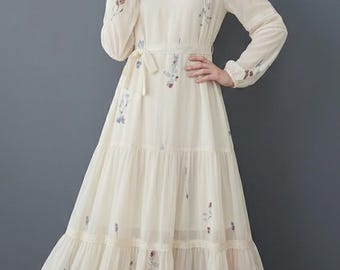 以下が含まれることがあります： クリーム色の長袖ドレス。ティアードスカートと花の刺繍が施されています。Vネック、レーストリム、お揃いのタイベルトが特徴です。ドレスは床までの長さで、流れるようなシルエットです。