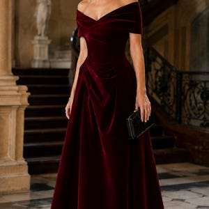 Puede incluir: Un vestido largo de terciopelo en color rojo burdeos con hombros descubiertos. El dise&ntilde;o presenta un escote cruzado, un corpi&ntilde;o ajustado con un sutil drapeado lateral y una falda larga. Se sostiene un peque&ntilde;o bolso de mano negro rectangular.