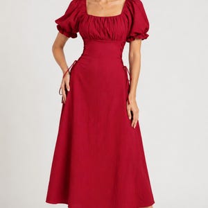 May include: A deep red midi dress featuring a square neckline, short puff sleeves with elasticated cuffs, and a fitted bodice with side lace-up corset detailing. The skirt flows into a relaxed A-line silhouette, crafted from a textured fabric.