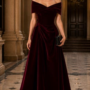 Puede incluir: Un vestido largo de terciopelo en color burdeos intenso con hombros descubiertos. El dise&ntilde;o presenta un escote cruzado, un corpi&ntilde;o ajustado y una falda drapeada con pliegues que llega hasta el suelo. Se sostiene un peque&ntilde;o bolso de mano negro.