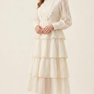 Può includere: Abito midi color crema con collo alto e maniche lunghe. L'abito presenta una gonna a balze e delicati dettagli in pizzo sul corpetto e sulle maniche. La modella indossa tacchi alti neri.