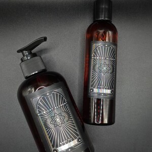 K&ouml;nnte beinhalten: Zwei dunkelbraune Glasflaschen mit Midnight Alchemy Body Oil. Eine Flasche hat einen Pumpspender, die andere einen schwarzen Deckel. Beide Flaschen haben ein schwarzes Etikett mit silbernen geometrischen Designs und dem Text "Midnight Alchemy" und "Body Oil".