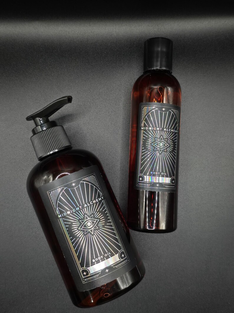 K&ouml;nnte beinhalten: Zwei dunkelbraune Glasflaschen mit Midnight Alchemy Body Oil. Eine Flasche hat einen Pumpspender, die andere einen schwarzen Deckel. Beide Flaschen haben ein schwarzes Etikett mit silbernen geometrischen Designs und dem Text "Midnight Alchemy" und "Body Oil".