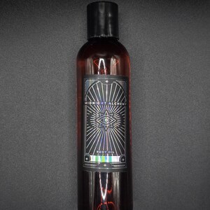 K&ouml;nnte beinhalten: Eine dunkel bernsteinfarbene K&ouml;rper&ouml;lflasche mit schwarzem Verschluss. Das Etikett tr&auml;gt die Aufschrift "MIDNIGHT ALCHEMY BODY OIL" mit einem Augensymbol und Strahlenlinien. Die Flasche steht vor dunklem Hintergrund.