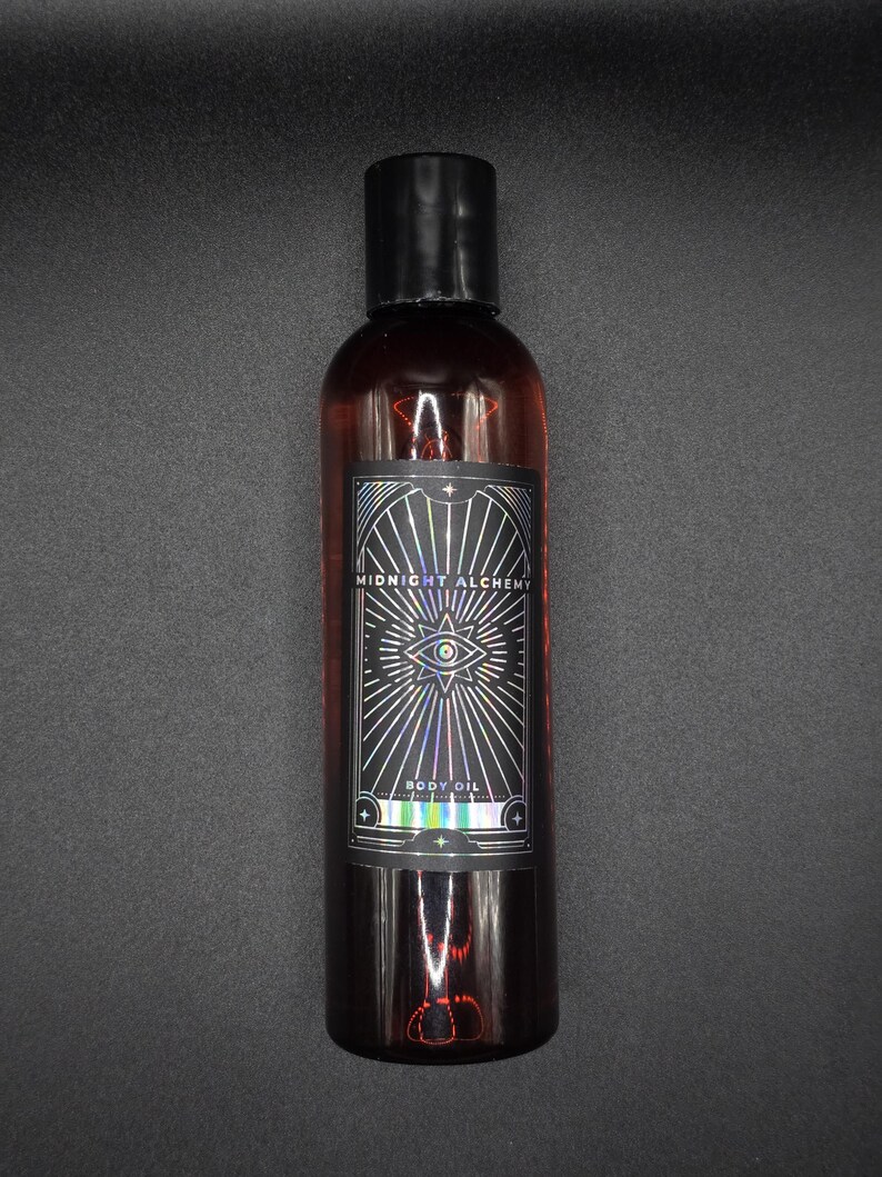 K&ouml;nnte beinhalten: Eine dunkel bernsteinfarbene K&ouml;rper&ouml;lflasche mit schwarzem Verschluss. Das Etikett tr&auml;gt die Aufschrift "MIDNIGHT ALCHEMY BODY OIL" mit einem Augensymbol und Strahlenlinien. Die Flasche steht vor dunklem Hintergrund.