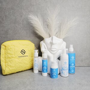 May include: A collection of skincare products, including bottles and a yellow quilted cosmetic bag, arranged on a gray surface. The bottles are white with blue labels and the text "Nanoestetic". A white face-shaped vase holds white decorative plumes.
