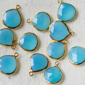 May include: A collection of light blue teardrop-shaped pendants with gold-colored edges and small loops for hanging. The pendants have a faceted surface, and are arranged on a neutral-colored fabric.