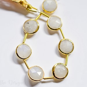 May include: Strand of round, faceted, pale gray moonstone beads with gold-colored metal settings and connecting wires. The beads are strung together, creating a delicate jewelry component. A gold tassel is visible at the top.