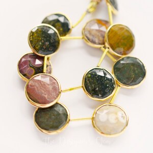 May include: Close-up of a strand of gemstone beads. The beads are round, with a faceted surface and a gold-colored metal rim. The gemstones are a mix of colors, including green, brown, and red. The beads are strung together with gold-colored wire.