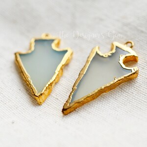 May include: Two arrowhead-shaped pendants with a pale blue center and gold-colored edges. The pendants are displayed on a white, textured surface. The text "The Designers Gem" is visible in the background.