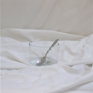 May include: A clear, cylindrical glass with a textured surface, containing a silver spoon. The spoon rests inside the glass, which is set against a white, draped fabric background. The glass appears to be empty.
