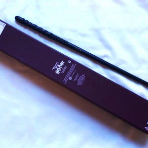 May include: A black wand with a textured handle lies next to a dark purple box. The box has the words "Harry Potter" printed on it. The wand is approximately 14 inches long.