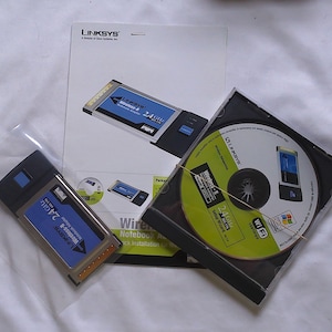 May include: A Linksys Wireless-B 2.4GHz notebook adapter, a CD-ROM, and a manual are displayed. The adapter is black and blue, with the text "Wireless-B" and "2.4GHz". The CD-ROM is in a black case, and the manual has the Linksys logo.