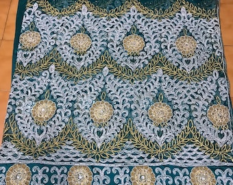 Puede incluir: Tela verde esmeralda con intrincados bordados blancos y dorados. El diseño presenta patrones florales y circulares, con un ribete de cuentas. La tela es probablemente para ropa formal o prendas de ocasión especial.