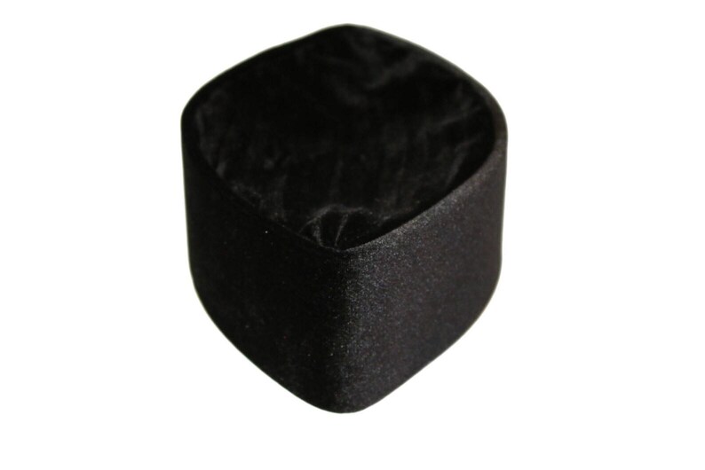Op de afbeelding: Een klein, vierkant zwart sieradendoosje met afgeronde hoeken. De buitenkant heeft een matte, getextureerde afwerking, terwijl de binnenkant is bekleed met een zachte, donkerzwarte stof die bij de basis licht geplooid of gerimpeld oogt.