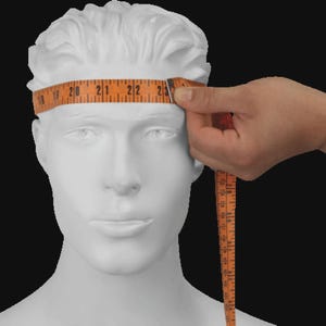 Op de afbeelding: Een wit etalagepophoofd met een gestructureerd kapsel. Een oranje meetlint is om het voorhoofd gewikkeld en toont een afmeting van ongeveer 58,4 cm. Het lint is voorzien van zwarte cijfers en lijnen voor een nauwkeurige hoofdomtrek.