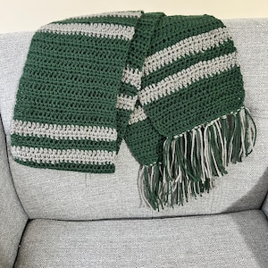 May include: A handmade, green and gray striped crochet scarf with fringe. The scarf is folded over a gray upholstered chair. The scarf is made of a soft yarn and has a textured appearance.