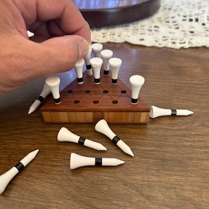 May include: A wooden peg solitaire game with a triangular board and white pegs with black bands. The board is made of dark wood with a lighter wood border. Some pegs are in the board, and others are scattered around.
