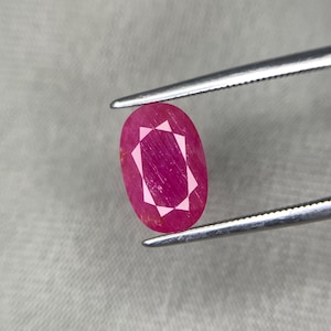 以下が含まれることがあります： 銀色のピンセットで保持された、楕円形のルビーの宝石。 ルビーは深い赤色で、内部にスターバーストパターンがあります。 宝石はニュートラルグレーの背景に配置されています。