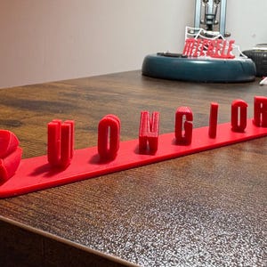 May include: A bright red 3D printed sign featuring the word BUONGIORNO in raised, block capital letters attached to a flat, elongated rectangular base. The letters are evenly spaced and stand vertically on the surface of the red plastic base.