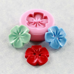 May include: A silicone mold for making flower-shaped buttons. The mold is pink and has a single cavity in the shape of a five-petal flower. Three finished buttons are shown, one green, one blue, and one red.