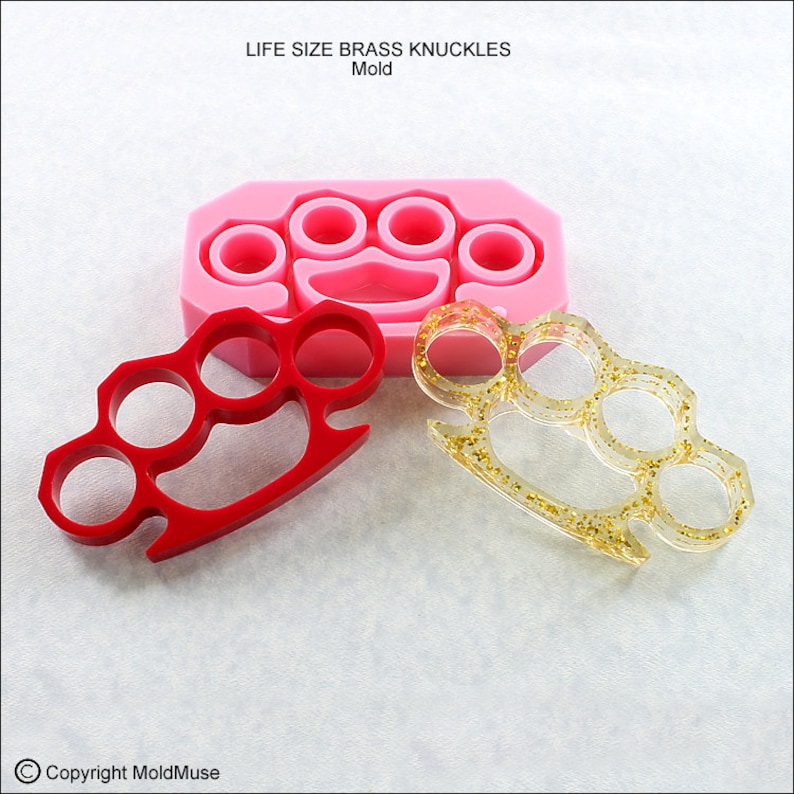 May include: A pink silicone mold for making brass knuckles. The mold is shown with two finished resin castings, one red and one clear with gold glitter.