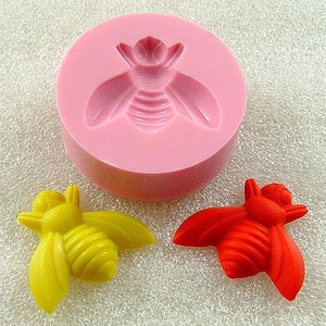 May include: A silicone mold for making bee-shaped decorations. The mold is pink and has a bee design in the center. There are two finished bee decorations, one yellow and one red, next to the mold.