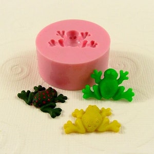 May include: Silicone mold for making frog-shaped decorations. The mold is pink and has a frog design. There are three finished frogs, one green, one yellow, and one green with red spots.