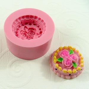 May include: Pink silicone mold for making a small cake with a detailed design of roses and leaves. The mold is shown with a finished cake made from it.