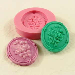 May include: A silicone mold for making resin or polymer clay cabochons. The mold is pink and has a design of a floral bouquet with a bow. There are two finished cabochons, one green and one pink, showing the design.