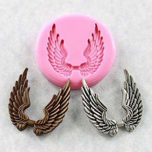May include: A pink silicone mold for making angel wings. The mold is shown with two finished wings, one in bronze and one in silver.