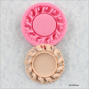 May include: A pink silicone mold with a detailed design of a round frame with a rope-like border and a decorative leaf pattern. The mold is shown next to a beige resin casting of the same design.