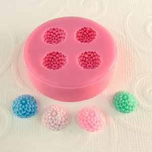 May include: A pink silicone mold with four flower-shaped cavities. Beside the mold are three flower-shaped objects in blue, pink, and green. The objects appear to be made of a solid material, possibly soap or resin, and are in the shape of a chrysanthemum.