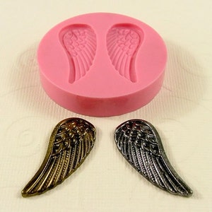 May include: A pink silicone mold with two angel wing shapes. Two finished angel wings are shown, one gold and one silver.