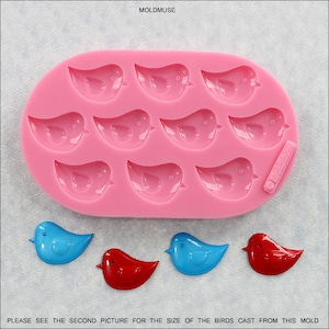 Può includere: Uno stampo in silicone rosa con 12 cavità a forma di uccello. Sotto lo stampo ci sono quattro oggetti finiti a forma di uccello in blu e rosso. Il testo "MOLDMUSE" è in alto e il testo "PLEASE SEE THE SECOND PICTURE FOR THE SIZE OF THE BIRDS CAST FROM THIS MOLD" è in basso.
