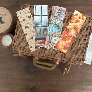 Könnte beinhalten: Ein Weidenkorb enthält illustrierte Lesezeichen mit Herbstmotiven, darunter Pilze, Bücher und Blätter. Eine Kerze und ein Behälter mit der Aufschrift "Natural Wood" sind ebenfalls vorhanden, was eine gemütliche, saisonale Atmosphäre schafft.