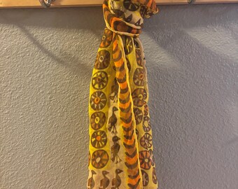 May include: A lightweight yellow scarf featuring a repeating pattern of brown and orange circular motifs, stylized birds, and crescent shapes. The fabric is semi-sheer with a signature printed near the bottom edge that reads Usna.