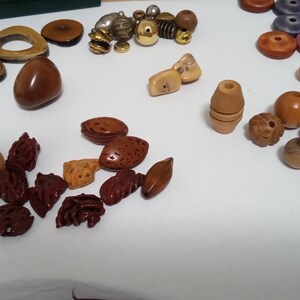 May include: A collection of various wooden beads in shades of brown, beige, and red, including round, oval, and carved shapes. Some beads have a glossy finish, while others are matte. There are also some metallic and multi-colored beads.