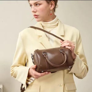 May include: A brown leather barrel bag with silver hardware, a top handle, and a detachable shoulder strap. The bag is held by a person wearing a yellow blazer.
