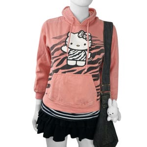 May include: A coral pink hoodie with a black and white zebra print design and a large Hello Kitty graphic. The hoodie has a front pocket, a black and white striped hem, and is paired with a black skirt and a denim shoulder bag with a white bunny charm.