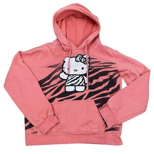May include: A coral pink hooded sweatshirt featuring a Hello Kitty graphic with a zebra print design. The graphic includes a white bow, a zebra-striped dress, and a waving hand. The sweatshirt has a kangaroo pocket and a drawstring hood.