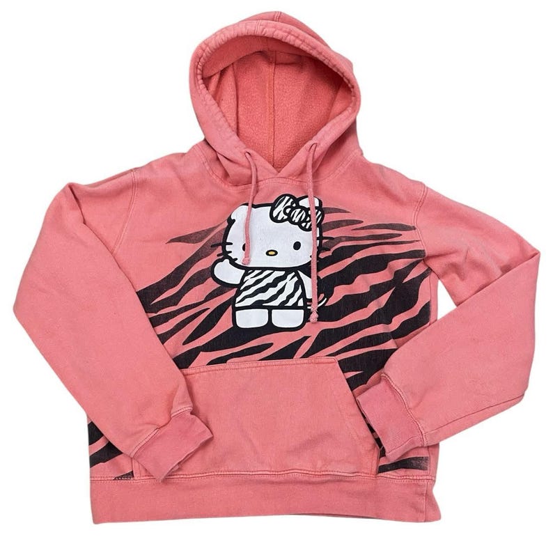 May include: A coral pink hooded sweatshirt featuring a Hello Kitty graphic with a zebra print design. The graphic includes a white bow, a zebra-striped dress, and a waving hand. The sweatshirt has a kangaroo pocket and a drawstring hood.