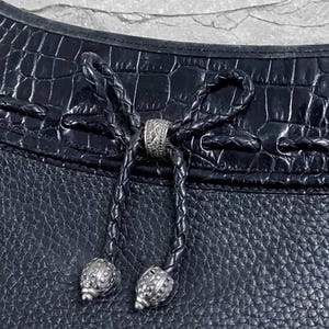 May include: Close-up of a black leather handbag with a textured surface. The bag features a braided leather drawstring closure with decorative silver-toned metal beads and a central ring. The top of the bag has a crocodile-like embossed pattern.