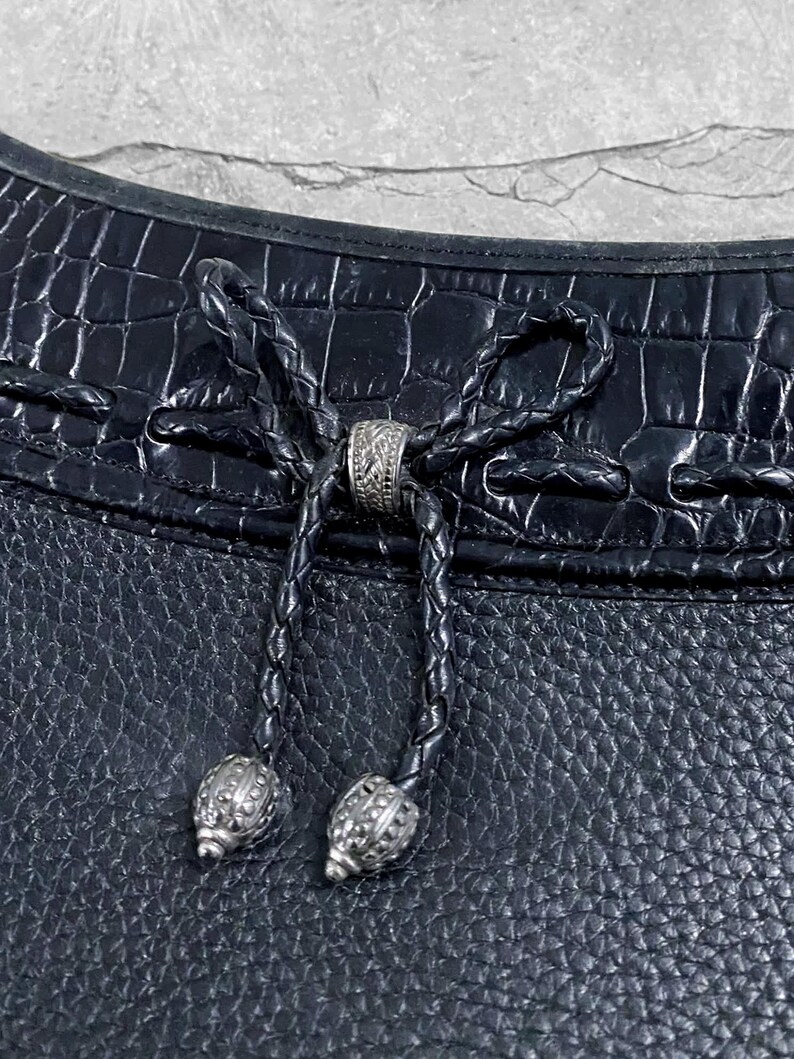 May include: Close-up of a black leather handbag with a textured surface. The bag features a braided leather drawstring closure with decorative silver-toned metal beads and a central ring. The top of the bag has a crocodile-like embossed pattern.