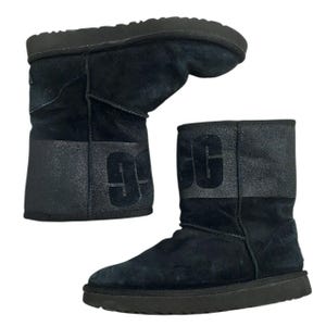 May include: Black suede boots with a dark gray band across the mid-section and the letters "UGG" in a dark gray color. The boots have a thick, black sole and are designed for cold weather.