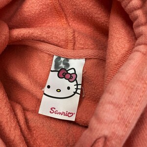 May include: Close-up of a coral-colored hoodie with a hood. The tag features the Hello Kitty character with a pink bow and the Sanrio logo in pink text.