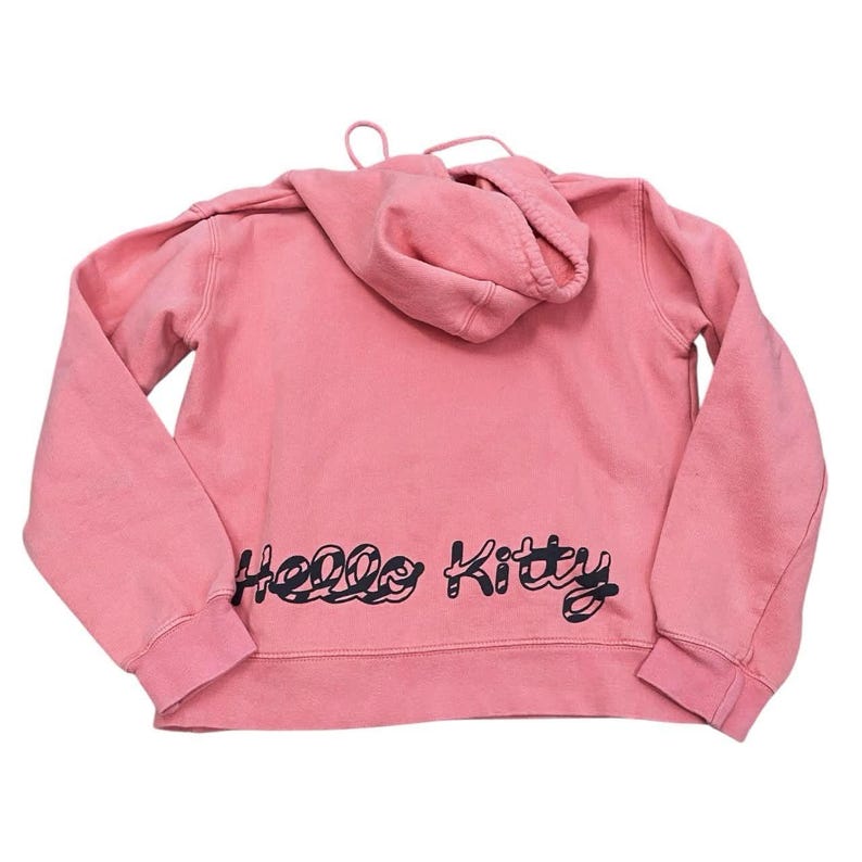 May include: A pink hooded sweatshirt with long sleeves. The back of the sweatshirt features the words "Hello Kitty" in black script. The hoodie has a drawstring and a kangaroo pocket.